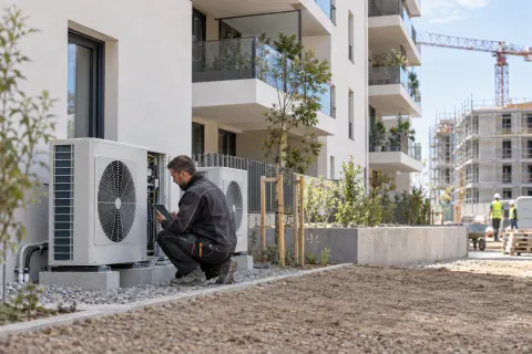 Technicien contrôlant une pompe à chaleur devant un immeuble de logements neufs en construction